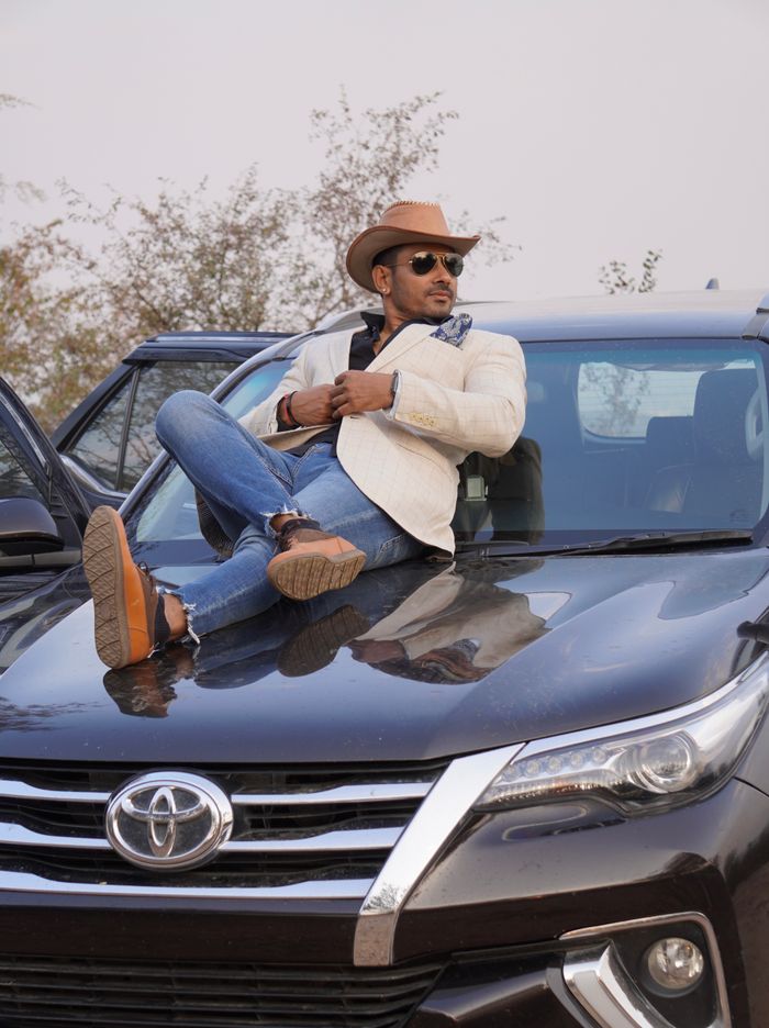 A fashionable man in a cowboy hat reclines on a Toyota SUV hood in an outdoor setting.